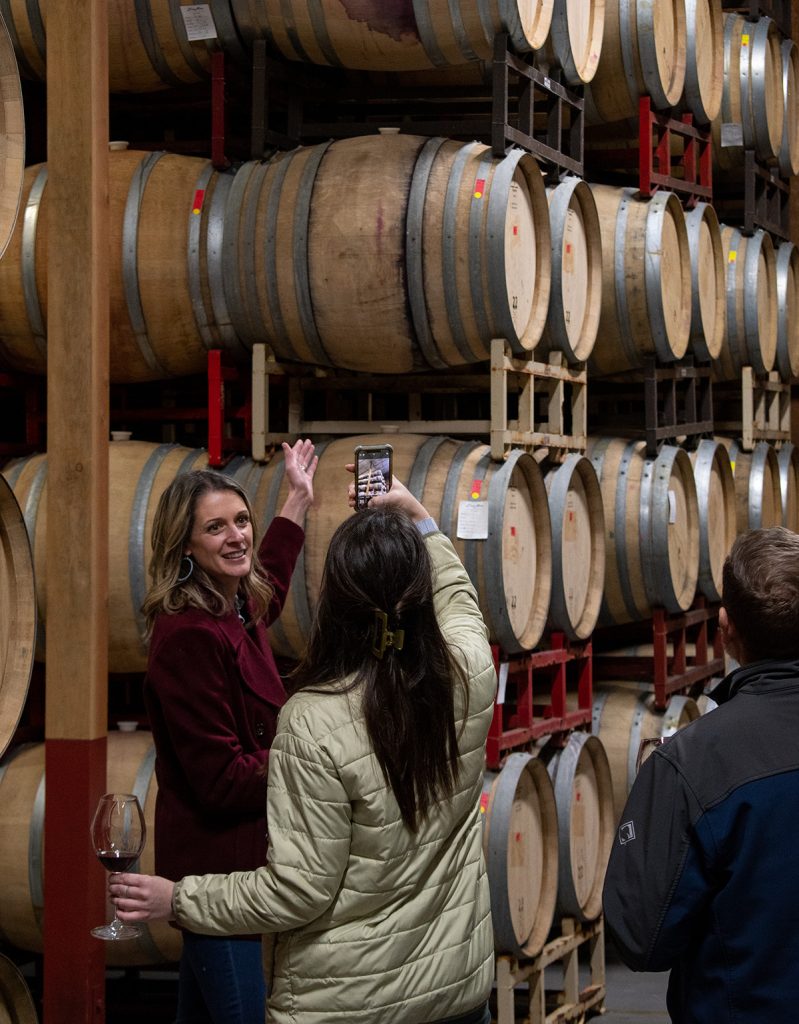 Couple on a private tour in the barrel room. A woman gestures to the barrels while another woman takes a photo.